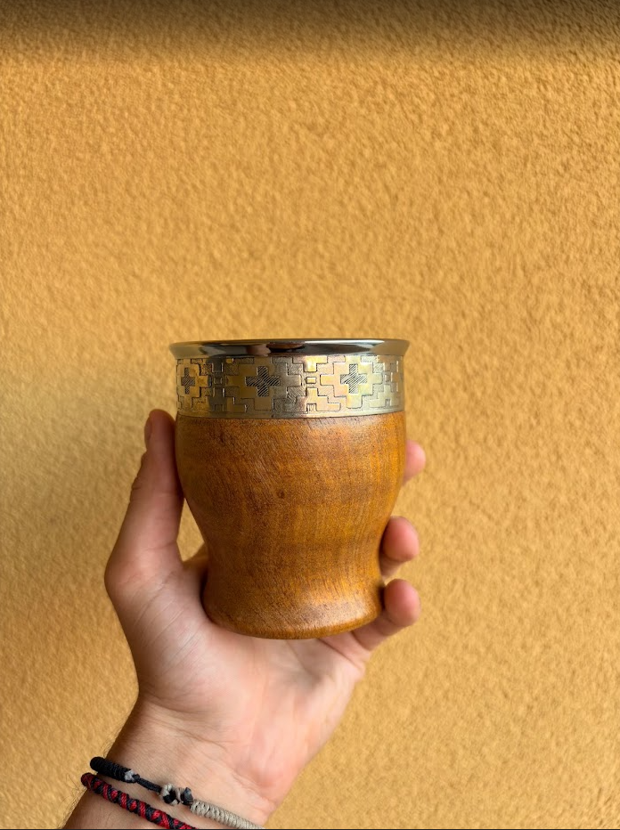 Mate de algarrobo de color madera natural con virola de alpaca grabada, sostenido en una mano.